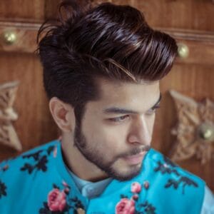 Close-up of a fashionable man with styled hair and beard wearing a floral vest indoors.