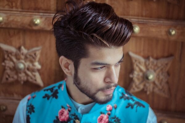 Close-up of a fashionable man with styled hair and beard wearing a floral vest indoors.